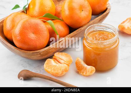 Krug mit gesunder Mandarinenkonfitüre und Holzschüssel mit Mandarinenorangen-Früchten auf weißem Küchentisch. Stockfoto