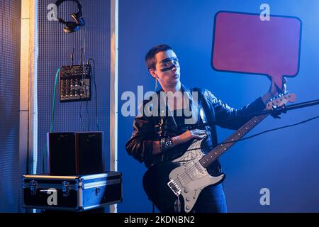 Rock-Künstler Mädchen hält Pappe Rede Blase und Gitarre, um Textnachricht auf isolierten Mockup Karton zu werben. Leere Plakatwand vor der Performance von Heavy Metal-Musik. Stockfoto