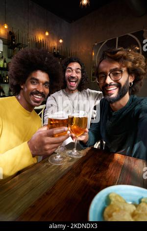 Vertikales Selfie von drei männlichen Freunden, die fröhlich in einer Bar toasten und mit großem Lächeln auf die Kamera blicken. Stockfoto