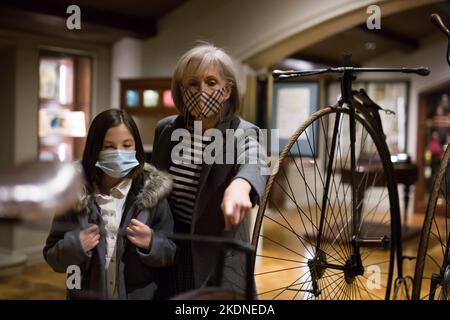 Zwischen einem Schulmädchen und einer älteren Tutorin in Gesichtsmasken beim Betrachten eines Vintage-Fahrrads im Museum Stockfoto