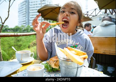 Das kleine Mädchen frühstückt in einem Café, isst Hamburger und pommes Frites, trinkt Schokolade Stockfoto