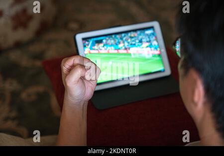 Mann mit der Faust nach oben, während er Fußballspiele auf dem digitalen Tablet auf der Couch ansieht. Stockfoto