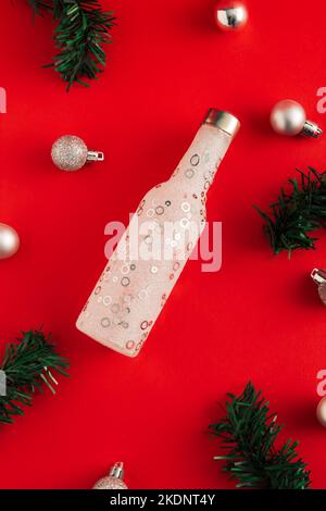 Neujahrskonzept aus Flasche, Tannenzweigen und Weihnachtskugeln auf rotem Hintergrund. Minimale Party-Idee. Flach liegend, Draufsicht. Stockfoto