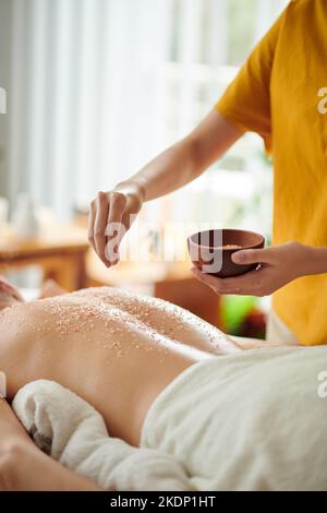 Zugeschnittenes Bild von Masseuse, die Salz schrubbt, auf dem Rücken der Frau Stockfoto