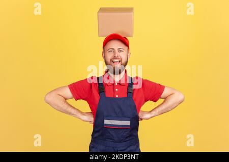 Porträt eines lustigen Kuriermanns in blauer Uniform, der mit den Händen auf den Hüften stand und den Karton auf dem Kopf hielt und lächelte auf die Kamera. Innenaufnahme des Studios isoliert auf gelbem Hintergrund. Stockfoto