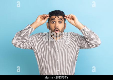 Überraschender Geschäftsmann, der mit erhobener Brille aufsteht, aufmerksam schaut, sieht etwas schockierendes und Erstauntes in einem gestreiften Hemd. Innenaufnahme des Studios isoliert auf blauem Hintergrund. Stockfoto