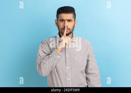 Bitte, Sei still. Geschäftsmann hält den Finger auf den Lippen und schweigt eine Geste, bittet um Geheimhaltung, spricht nicht, trägt gestreiftes Hemd. Innenaufnahme des Studios isoliert auf blauem Hintergrund. Stockfoto