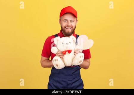 Porträt eines aufgeregten glücklichen Kuriermanns in blauer Uniform und roter Mütze, der weichen Teddybär und Herz auf dem Stock hält und die Kamera anschaut. Innenaufnahme des Studios isoliert auf gelbem Hintergrund. Stockfoto