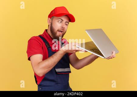 Seitenansicht Porträt eines bärtigen Arbeiters in blauer Uniform und roter Mütze, der ein halb geöffnetes Notizbuch hält und verbotene Informationen ausspioniert. Innenaufnahme des Studios isoliert auf gelbem Hintergrund. Stockfoto
