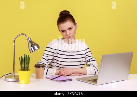 Gelangweilte, unglückliche Büromanagerin mit gestreiftem Hemd, die am Arbeitsplatz sitzt und die Kamera anschaut, müde von Online-Konferenzen. Studio-Innenaufnahme isoliert auf gelbem Hintergrund. Stockfoto