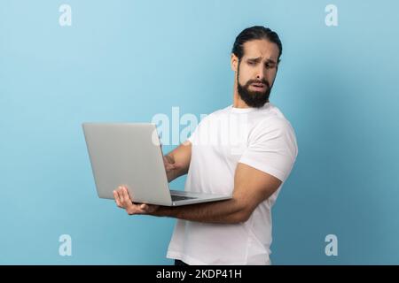 Porträt eines gutaussehenden Mannes mit Bart, das weißes T-Shirt trägt, das sein Gesicht vom Display abdreht, weil er nicht auf dem Laptop-Bildschirm sehen möchte. Innenaufnahme des Studios isoliert auf blauem Hintergrund. Stockfoto