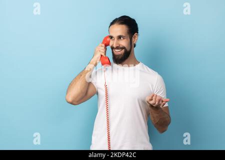 Porträt eines positiven Mannes mit Bart in weißem T-Shirt, der auf einem roten Retro-Telefon spricht, angenehme Gespräche führt, mit einem Lächeln wegschaut Innenaufnahme des Studios isoliert auf blauem Hintergrund. Stockfoto