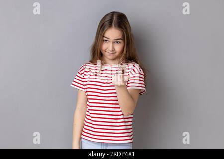 Porträt eines kleinen Mädchens in gestreiftem T-Shirt, das spielerisch aussieht und mit einem Finger ruft, was eine winkende Geste macht, die zum Kommen einlädt. Innenaufnahme des Studios isoliert auf grauem Hintergrund. Stockfoto