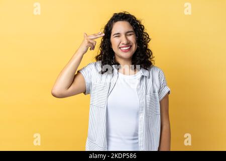 Porträt einer Frau mit dunklen welligen Haaren, die mit der Fingerpistole auf den Kopf zeigt und verzweifelt aussieht, Selbstmordgesten macht und sich selbst schießt. Innenaufnahme des Studios isoliert auf gelbem Hintergrund. Stockfoto