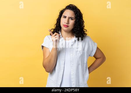 Porträt einer ernsthaften strengen Frau mit dunklen welligen Haaren, die mit dem Finger auf die Kamera zeigt und mit unzufriedenem, verdächtigem Ausdruck aussieht. Innenaufnahme des Studios isoliert auf gelbem Hintergrund. Stockfoto