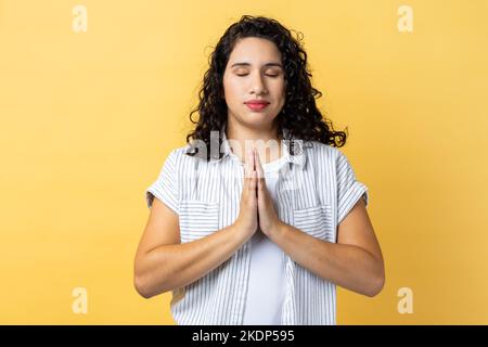 Porträt einer friedlichen, ruhigen, entspannten Frau mit dunklen, welligen Haaren, die mit Gebetstus steht, die Augen geschlossen hält und in Yoga-Position meditiert. Innenaufnahme des Studios isoliert auf gelbem Hintergrund. Stockfoto