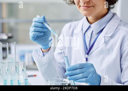 Junge Laborarbeiterin in Handschuhen und Laborkittel, die während wissenschaftlicher Untersuchungen flüssige Substanz von der Pipette in den Kolben tropft Stockfoto