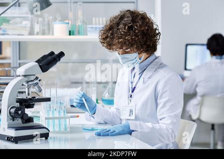 Junger Biochemiker in schützender Arbeitskleidung, der sich während des wissenschaftlichen Experiments über den Schreibtisch beugt, während er zwei flüssige Substanzen in einer Petrischale mischt Stockfoto