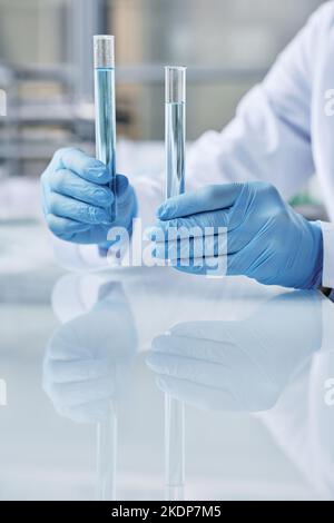 Handschuhen junger Virologen, die zwei Flaschen mit Flüssigkeiten halten, die während des Experiments im wissenschaftlichen oder klinischen Labor hergestellt wurden Stockfoto