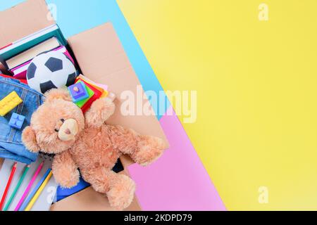 Spendenbox mit Kinderspielzeug, Büchern, Kleidung für wohltätige Zwecke auf buntem Gelb, Rosa, hellblauem Hintergrund. Draufsicht Stockfoto
