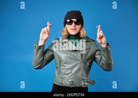 Hübsche junge blonde Frau in grüner Jacke posiert isoliert auf blauem Hintergrund. Erstellen Sie ein Modell für den Kopierbereich. Auf einen besonderen Moment warten, die Daumen drücken, Stockfoto