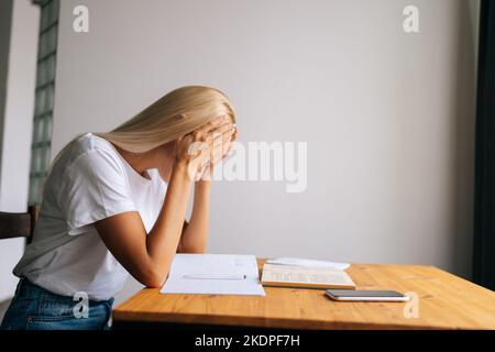 Seitenansicht der müden blonden jungen Frau, die Tempel massierte, nach dem Papierkram Kopfschmerzen litt, zu Hause mit Buch und Notizbuch am Schreibtisch saß. Stockfoto