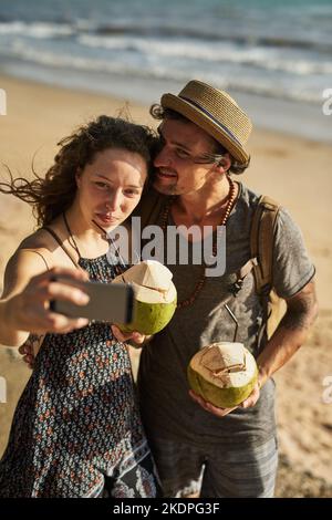 Unsere Freunde werden so eifersüchtig sein. Ein glückliches junges Paar, das Selfies macht, während es Cocktails am Strand genießt. Stockfoto