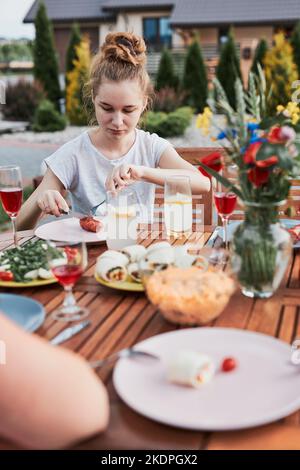 Junge Frau beim Essen vom Grill während des Sommerpicknicks für die Familie im Freien in einem Hausgarten. Nahaufnahme des Tisches mit Speisen und Geschirr Stockfoto