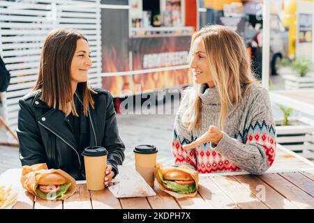 Zwei glückliche hungrige Freundinnen plaudern und essen Burger auf dem Straßenmarkt. Stockfoto