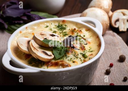 Hühnerjulienne mit Käse, Pilzen und Kräutern in weißem Teller auf Holzbrett Stockfoto