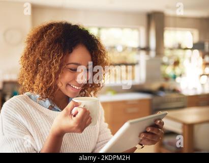 Es ist Zeit, sich zu vernetzen und die neuesten Online-Informationen zu erhalten. Eine glückliche junge Frau, die ein digitales Tablet nutzt und zu Hause in der Küche Kaffee trinkt. Stockfoto