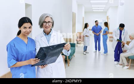 Weibliche Praktikantin mit ihrem medizinischen Mentor im Krankenhauskorridor während der medizinischen Praxis vor dem Hintergrund von Ärzten und medizinischem Personal. Praktikum und Sanitäter Stockfoto