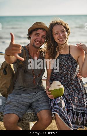 Entspannt und liebend. Porträt eines glücklichen jungen Paares, das Cocktails genießt und sich auf einer Bank am Strand entspannt. Stockfoto