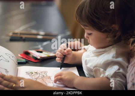 Ein junger, entzückender Kleinkind saß mit seiner Mutter und zeichnete mit Buntstiften in einem eingefärbten Buch Stockfoto