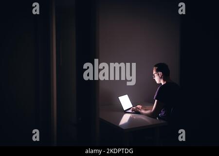 Nachtarbeit mit Laptop in dunklem Home Office-Zimmer. Junger Mann mit Notebook-Computer. Coder, Programmierer, Hacker, freiberuflicher digitaler Redakteur oder Student. Stockfoto