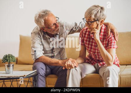 Seniorenpaar sitzt im Wohnzimmer. Die Frau ist traurig und besorgt und der Mann tröstet sie. Stockfoto