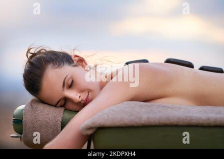 Eine junge Frau, die draußen auf einem Massageliegen eine Behandlung mit heißen Steinen erhält. Stockfoto