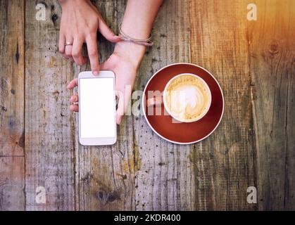 Anschluss über einen cremigen Cappuccino. Aufnahme einer Frau, die ihr Handy benutzt und eine Tasse Kaffee trinkt. Stockfoto