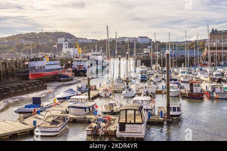 Schiffe und Yachten teilen sich einen Hafen mit einem Leuchtturm am Ende eines Piers. Weiße Gebäude befinden sich auf der fernen Skyline und ein wolkiger Himmel ist darüber. Stockfoto