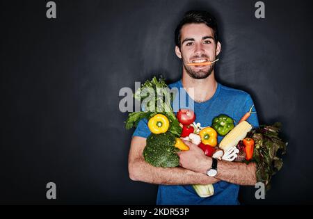 Essen Sie gut, sehen Sie gut aus, fühlen Sie sich gut. Studioporträt eines jungen Mannes, der vor dunklem Hintergrund eine Armvoll gesundem Gemüse trägt. Stockfoto