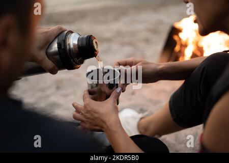 Ein paar Reisende trinken Kaffee. Herbstpicknick am Meeresstrand. Mann gießt Tee aus Thermoskannen. Frau hält Metallbecher mit Getränk. Lifestyle-Moment bei Stockfoto