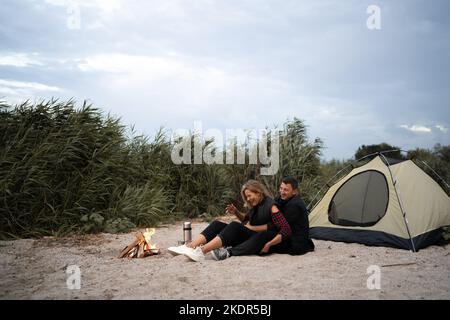 Romantische Touristen lachen am Lagerfeuer in der Nähe des Zeltes und umarmen sich unter Bäumen und Nachthimmel. Nachtcamping. Sorgloses Paar entspannend Stockfoto