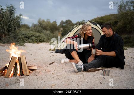 Ein fröhliches Paar sitzt in der Nähe eines Zeltes am Lagerfeuer und gießt am Wochenende Kaffee oder Tee aus einer Thermoskanne an einer Küste. Camping, Erholung Stockfoto