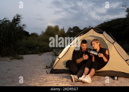 Nachtcamping am Strand. Mann und Frau Touristen ruhen sich vor einem Zelt unter dem Abendhimmel aus, trinken Kaffee am Strand und Stockfoto