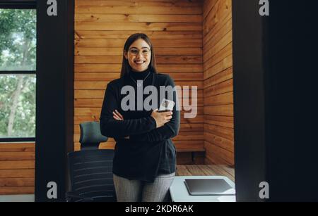 Erfolgreiche junge Geschäftsfrau lächelt an der Kamera, während sie ein Smartphone hält. Fröhliche Unternehmerin, die in einem modernen Co-Working aus der Ferne arbeitet Stockfoto