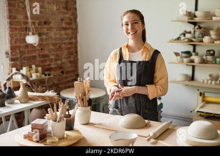 Waist-up-Porträt einer lächelnden jungen Frau mit Schürze in gemütlichem Töpferstudio, Kopierraum Stockfoto