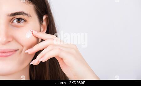 Nahaufnahme einer jungen Frau, die weichmachende Gesichtscreme anwendet, lächelnd zufrieden auf die Kamera, isoliert auf weißem Hintergrund mit Kopierraum. Hautpflegekonzept, Spa PR Stockfoto