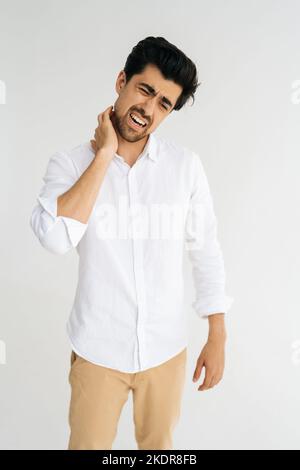 Vertikales Porträt eines müden bärtigen Mannes, der sich körperlich unwohl fühlt und angespannte Muskeln massiert, schmerzhaften Hals auf weißem isoliertem Hintergrund hat. Stockfoto
