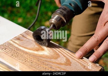 Die männliche Hand hält die Bürstenmaschine elektrisch rotierend, wobei die Metallscheibe ein Stück Holz schleift. Stockfoto