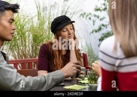 Nachdenkliche junge Rothaarige Frau, die mit Freunden Latte im Garten der Kaffeebar trinkt - glückliche Freunde, die im Hostel Dehors but thei miteinander reden und Spaß haben Stockfoto